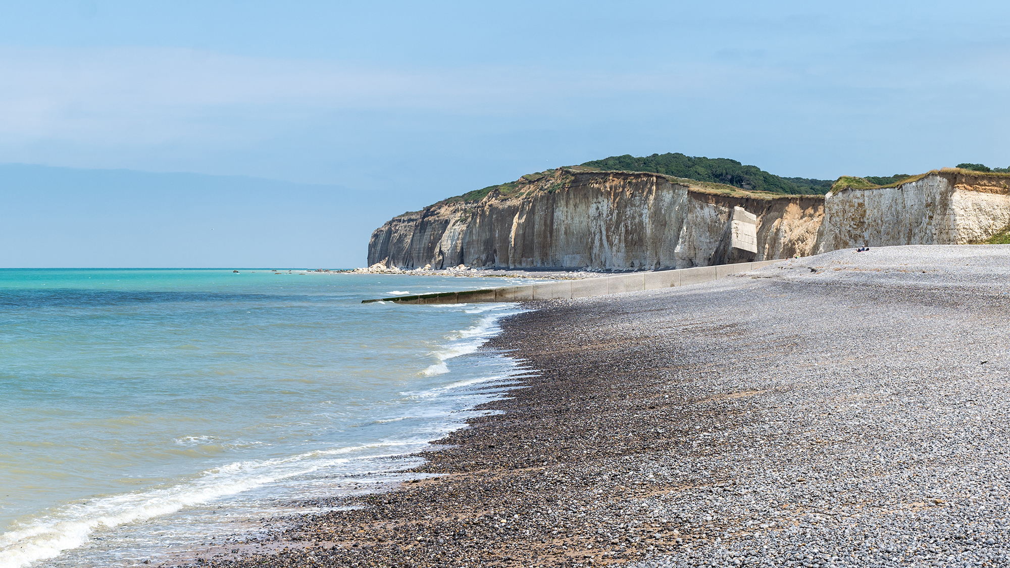 Normandy pebbled beach with clear blue waves washing onto the shore with green cliff tops in the background